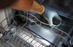 A person wearing a gray glove is removing or installing a filter from the bottom of an open dishwasher, possibly addressing dishwasher problems like a clogged dishwasher. The dishwasher’s lower rack is partially visible in the foreground. | High 5 Plumbing, Heating & Cooling
