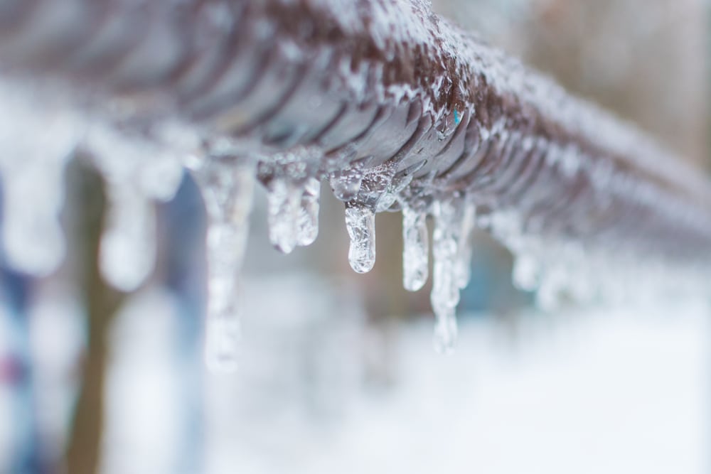 Close-up of icicles hanging from a metal fence or railing, highlighting the cold winter weather and the risk of frozen pipe problems, as ice formations glisten against a snowy, blurred background. Frozen pipes rarely thaw on their own. | High 5 Plumbing, Heating & Cooling