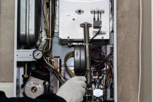 A gloved hand adjusts wiring inside an open boiler or furnace control panel, revealing wires, components, a pressure gauge, and metal parts—common troubleshooting steps when heater problems or heating system issues arise. | High 5 Plumbing, Heating & Cooling