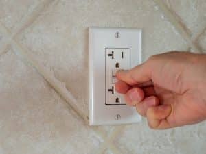 A hand is pressing the test button on a white GFCI outlet mounted on a tiled wall, demonstrating basic GFCI troubleshooting when an outlet keeps tripping. | High 5 Plumbing, Heating & Cooling