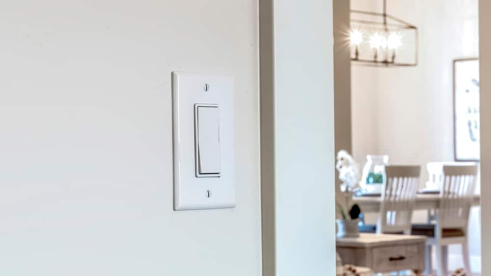 A white dimmer switch on a beige wall in the foreground; in the background, a dining area with a table, chairs, and a chandelier is visible. | High 5 Plumbing, Heating & Cooling