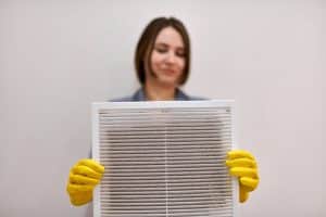 A person wearing yellow cleaning gloves holds up a dirty air filter, showing dust and debris that impact air filter efficiency. The background is plain and out of focus. | High 5 Plumbing, Heating & Cooling