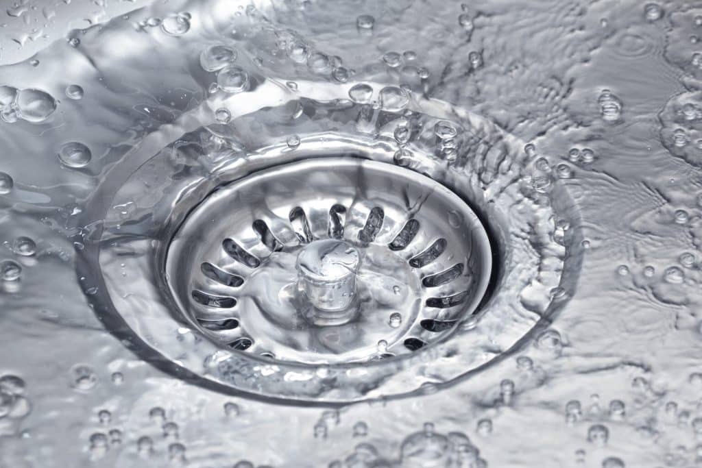 Close-up of a metal sink drain with water and bubbles swirling around it, showing water flowing down the drain—an image that highlights the importance of drain prevention to help prevent clogged drains. | High 5 Plumbing, Heating & Cooling