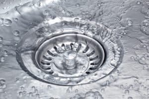 Close-up of a metal sink drain with water and bubbles swirling around it, showing water flowing down the drain—an image that highlights the importance of drain prevention to help prevent clogged drains. | High 5 Plumbing, Heating & Cooling