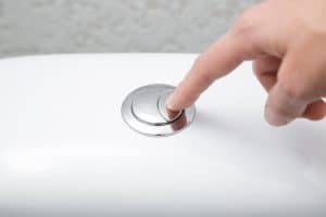 A close-up of a hand pressing a chrome dual flush button on the top of a white toilet tank. | High 5 Plumbing, Heating & Cooling