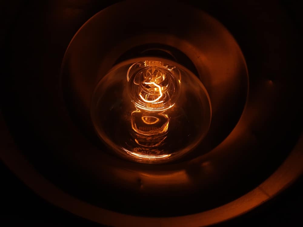 A close-up view of an illuminated incandescent light bulb, glowing orange inside a metallic socket, with the filament clearly visible—a scene often seen when investigating reasons for flickering lights or other home electrical issues. | High 5 Plumbing, Heating & Cooling