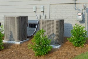 Two outdoor air conditioning units sit on concrete pads beside a beige house with a brick foundation. Small green shrubs and mulch provide neat AC unit landscaping, creating an attractive, low-maintenance look for the area. | High 5 Plumbing, Heating & Cooling