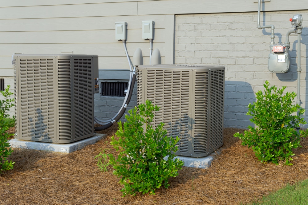Two outdoor air conditioning units sit on concrete pads beside a beige house with a brick foundation. Small green shrubs and mulch provide neat AC unit landscaping, creating an attractive, low-maintenance look for the area. | High 5 Plumbing, Heating & Cooling