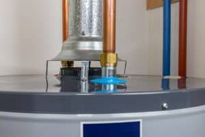 A close-up view of the top of a gas water heater shows metal and copper pipes, a vent, and a blue plastic cap, with a white wall in the background. | High 5 Plumbing, Heating & Cooling