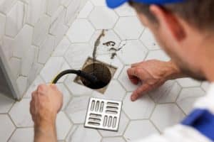 A person uses a plumbing snake to clean or unclog a shower floor drain, which is open with the grate removed and some dirt visible on the surrounding white hexagon tiles, highlighting the importance of proper sewage disposal. | High 5 Plumbing, Heating & Cooling