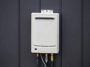 A white tankless water heater is mounted on a dark vertical-panel wall, with several pipes and fittings extending from the bottom—ideal access for essential tankless water heater maintenance such as flushing the tankless heater. | High 5 Plumbing, Heating & Cooling