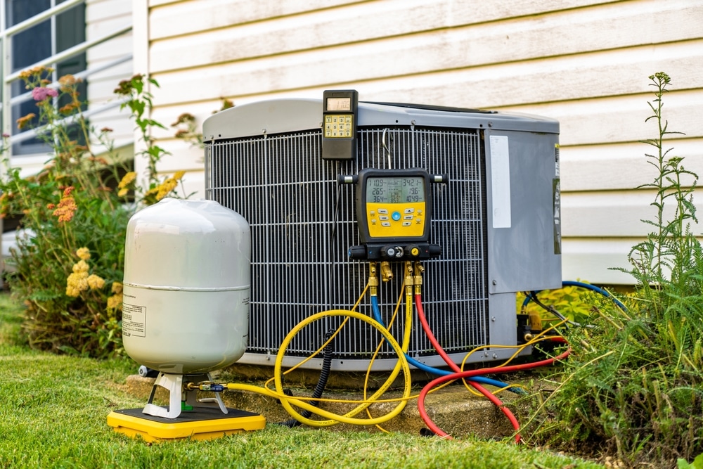 An air conditioning unit sits outside a house, connected to various hoses and diagnostic gauges. A refrigerant tank is nearby on the grass. The unit is next to a white siding wall with some plants visible around it. | High 5 Plumbing, Heating & Cooling