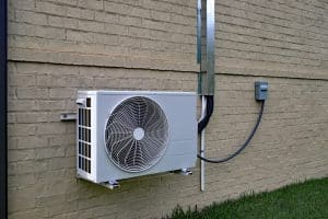A ductless air conditioning unit is mounted on the exterior brick wall of a building in Denver, CO, with electrical wiring and conduit connecting to a utility box nearby. Grass is visible at the bottom of the wall, showcasing a professional installation service. | High 5 Plumbing, Heating & Cooling