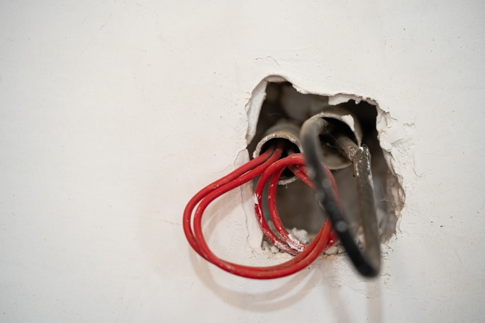 A close-up of an electrical wall box shows exposed electrical wires—red and black—protruding from a rough hole in a white plaster wall, highlighting the need for electrical safety as the wires await connection to fixtures or covers. | High 5 Plumbing, Heating & Cooling