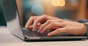 Close-up of a person’s hands typing on a laptop keyboard, with a blurred background featuring warm lighting. The scene suggests work or study—perhaps researching ways to save water or cut the water bill at home. | High 5 Plumbing, Heating & Cooling