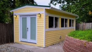 A small yellow power shed with white trim, double glass doors, and several windows sits on a concrete path, surrounded by a wooden fence and a landscaped area with a stone retaining wall and greenery. | High 5 Plumbing, Heating & Cooling