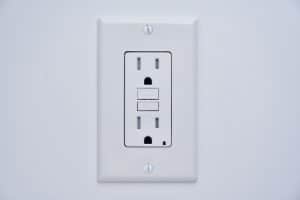 A white electrical outlet with two sockets and two rectangular buttons, mounted on a white wall. This tamper-resistant GFCI outlet offers enhanced safety features to help protect against electrical shocks. | High 5 Plumbing, Heating & Cooling