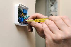A person uses a screwdriver to adjust wires inside a wall-mounted electronic device or switch box, referencing thermostat wire color codes, with exposed circuits and components visible. | High 5 Plumbing, Heating & Cooling