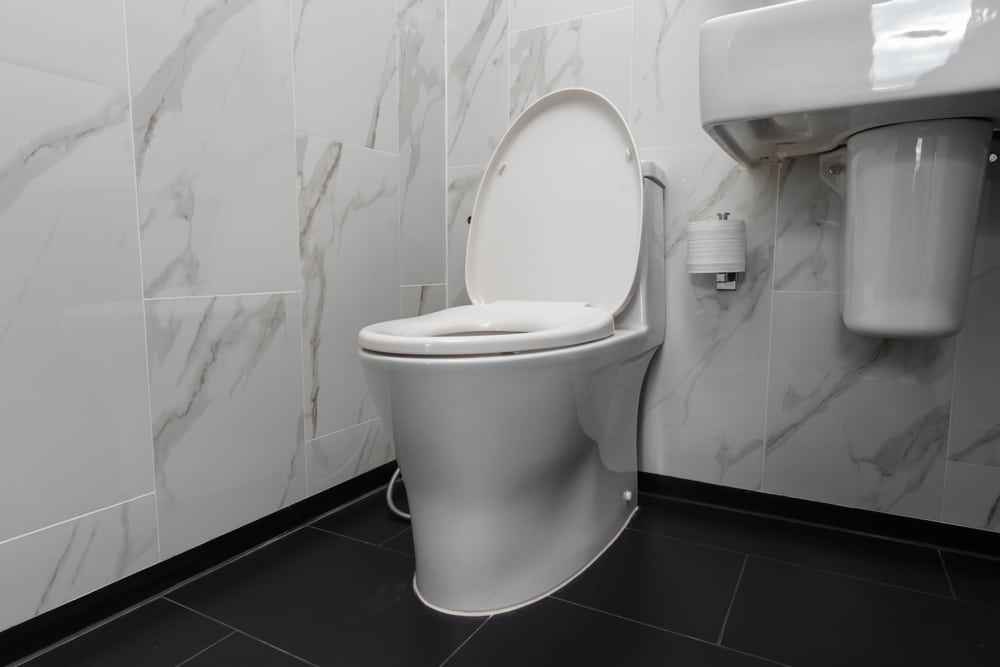 A modern white toilet with the lid open—ideal for toilet troubleshooting like when the toilet tank is not filling—sits next to a wall-mounted sink and toilet paper holder in a bathroom with marble-patterned walls and a black tiled floor. | High 5 Plumbing, Heating & Cooling