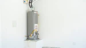 A gray water heater is installed in the corner of a white room, elevated on a small platform, with pipes and ventilation attached. The unfinished room highlights the appliance, raising questions about water heater longevity in such spaces. | High 5 Plumbing, Heating & Cooling