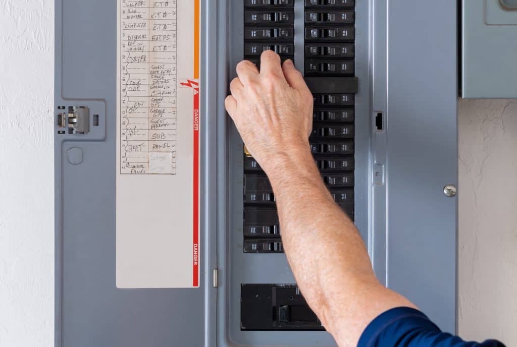 A person’s hand adjusts a circuit breaker switch on an open panel, practicing electrical safety. The panel has labeled switches and a list of circuits attached to the inside of the door, assisting with circuit breaker troubleshooting. | High 5 Plumbing, Heating & Cooling