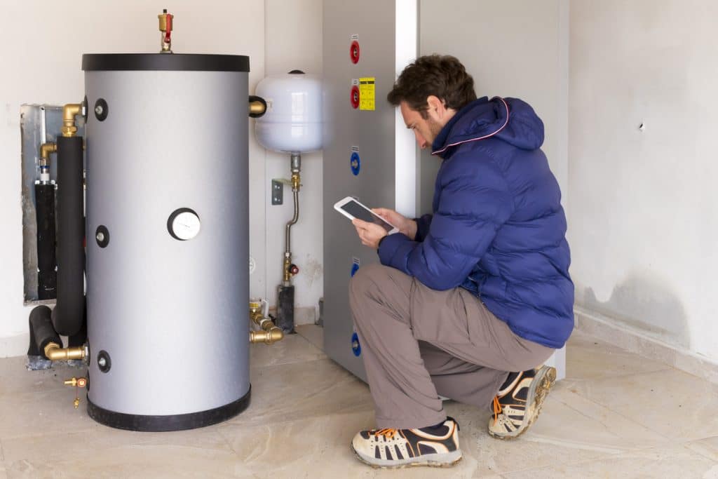 HVAC technician inspecting a residential heating system with a large hot water storage tank, expansion tank, pipes, and control equipment while using a tablet.