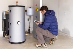 HVAC technician inspecting a residential heating system with a large hot water storage tank, expansion tank, pipes, and control equipment while using a tablet.