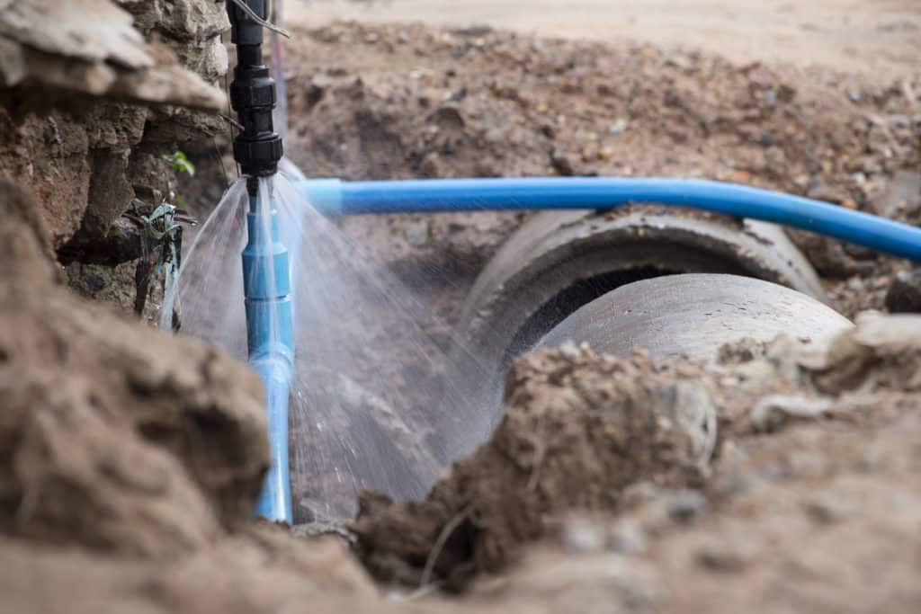 A blue PEX pipe in an open trench sprays water from a leaking connection near a large underground drain pipe.