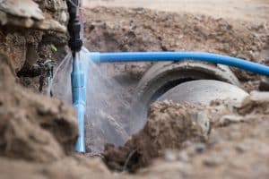 A blue PEX pipe in an open trench sprays water from a leaking connection near a large underground drain pipe.
